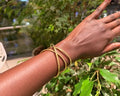 Suswa Bangle – handcrafted brass crossover wrap bangle worn on wrist against green foliage
