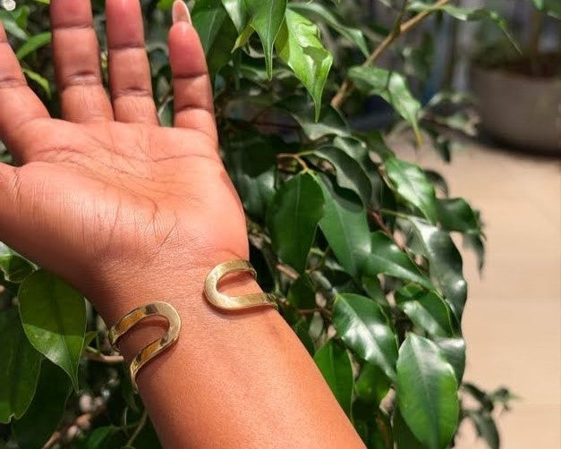 Ewaso Bangle – handcrafted brass horseshoe open cuff worn on wrist against green foliage