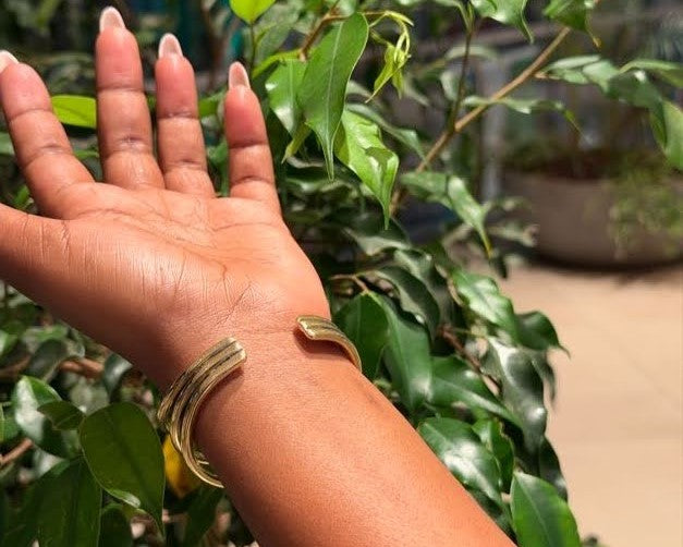 Ololua Bangle – handcrafted brass open wave cuff worn on wrist against green foliage